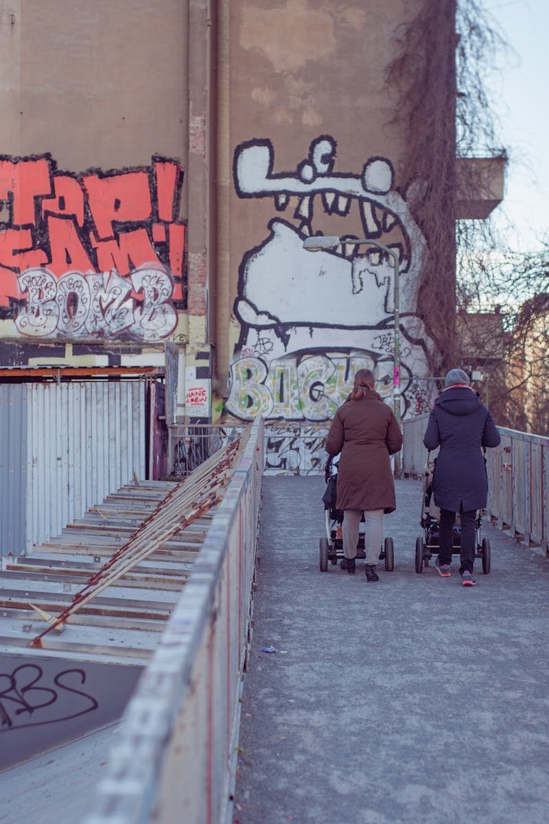 Mothers walking with strollers on graffiti-lined urban street bridge.