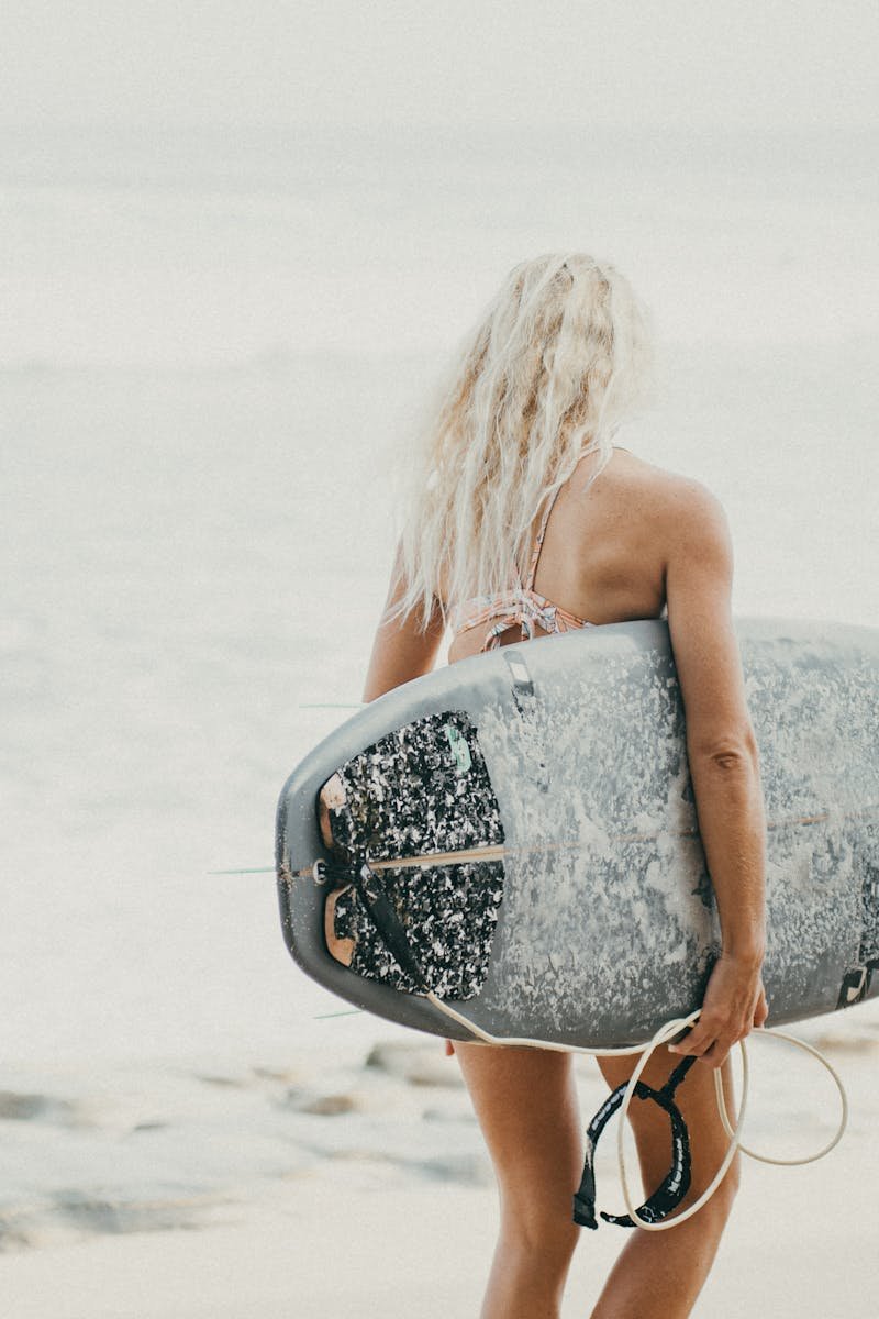 An adult woman carrying a surfboard walks towards the ocean on a sunny day in Bali, Indonesia.