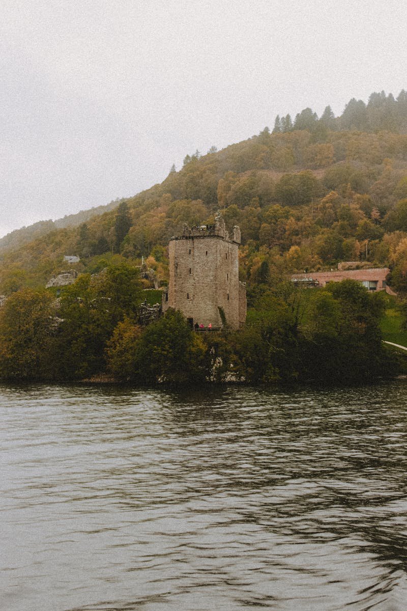 Scenery view of old tower facade on mount with trees near rippled lake under white sky in Scotland