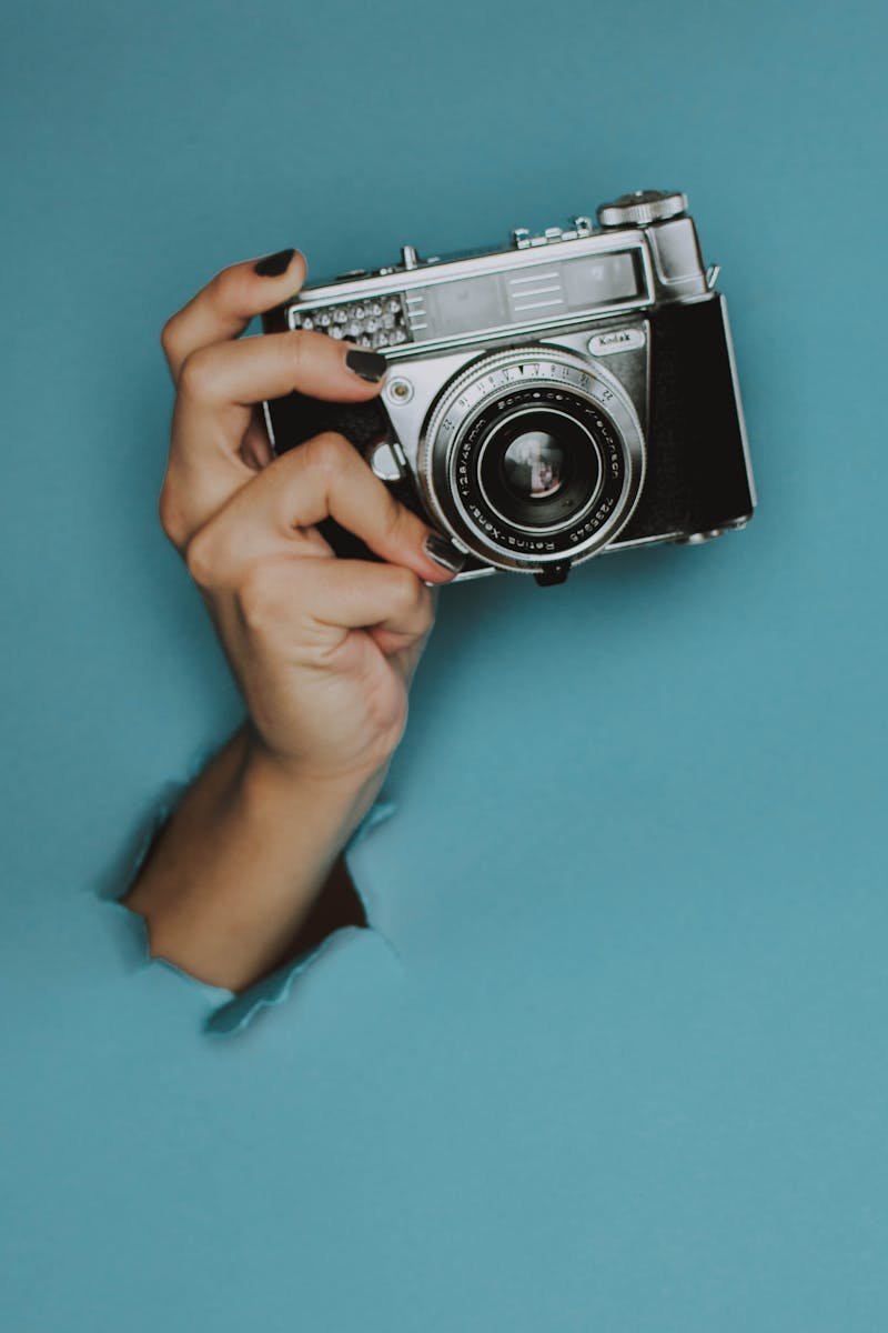A hand holds a classic vintage camera through a torn blue paper background.