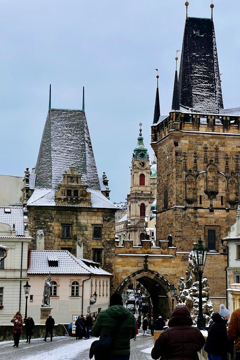Snow-covered towers at Charles Bridge with people strolling, capturing Prague's historic charm.